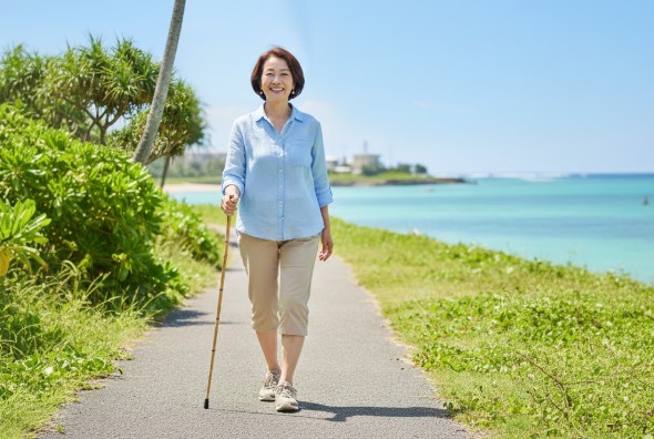 沖縄の海沿いの道を、杖を使いながらも笑顔で軽やかに散歩する60代の女性。うるま市の専門整体で股関節の痛みを根本改善し、歩く楽しみを取り戻した様子。
